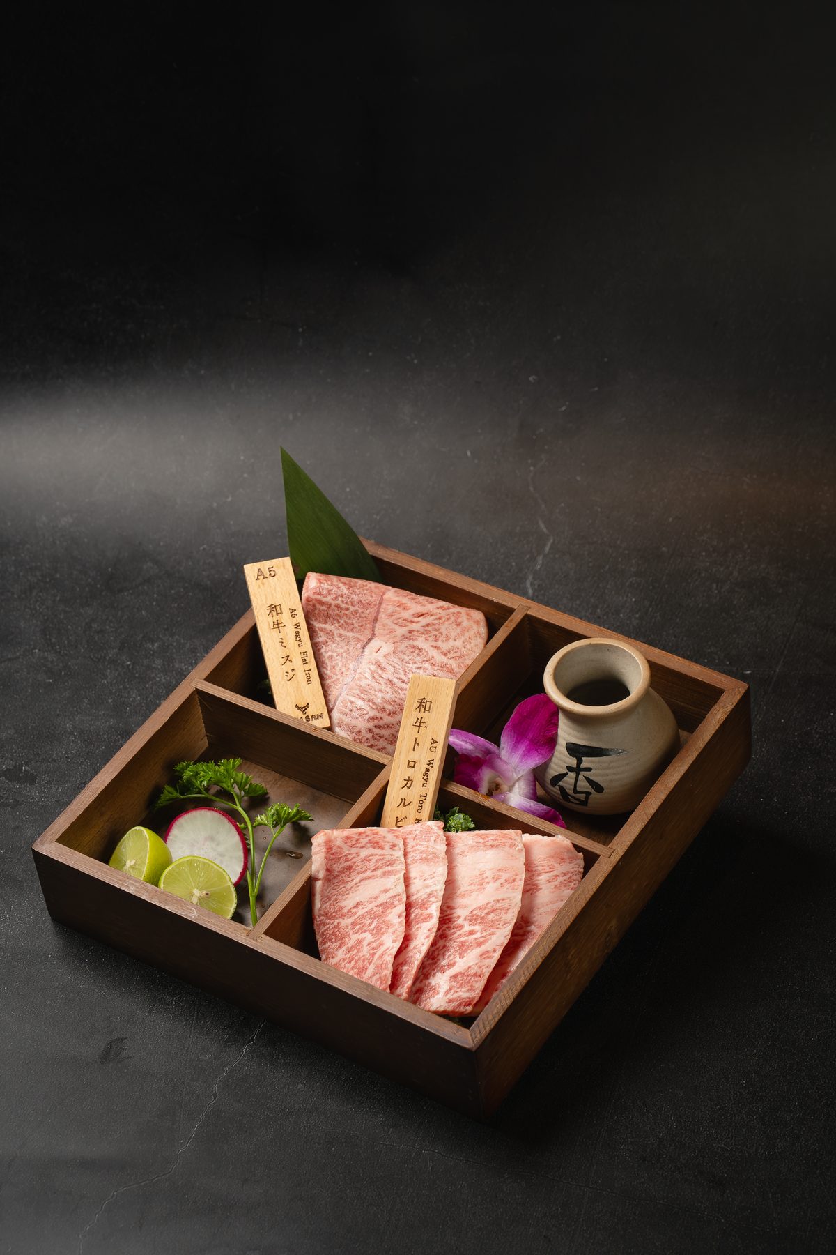 A wooden box with two portions of marbled A5 Wagyu, tagged in Japanese, with an orchid and lime slices at Gyusan