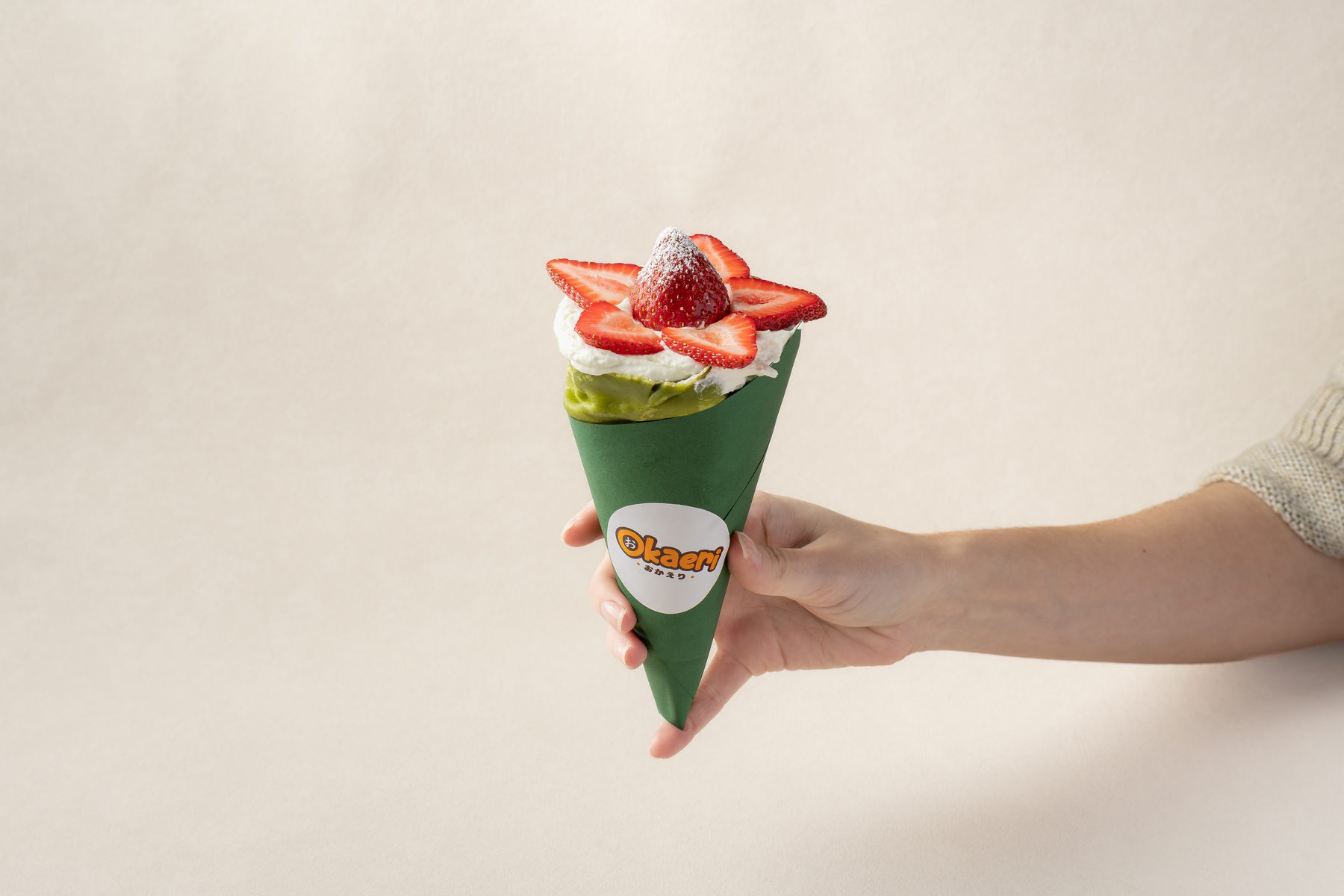 A hand holding a matcha-green crepe cone filled with cream and sliced strawberries from Okaeri in Centreville